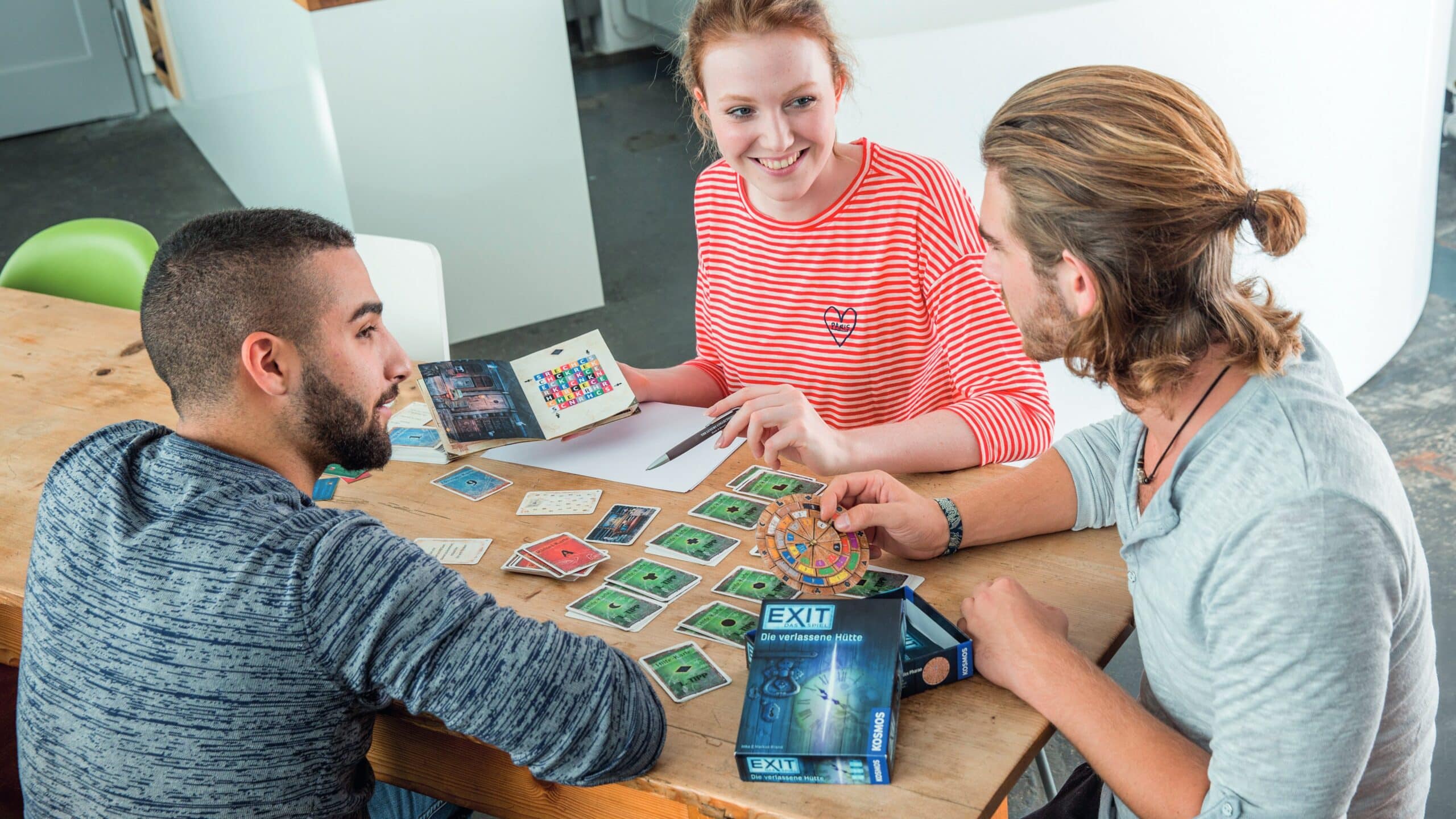 Trois amis sont assis à une table et jouent au jeu EXIT La cabane abandonnée.